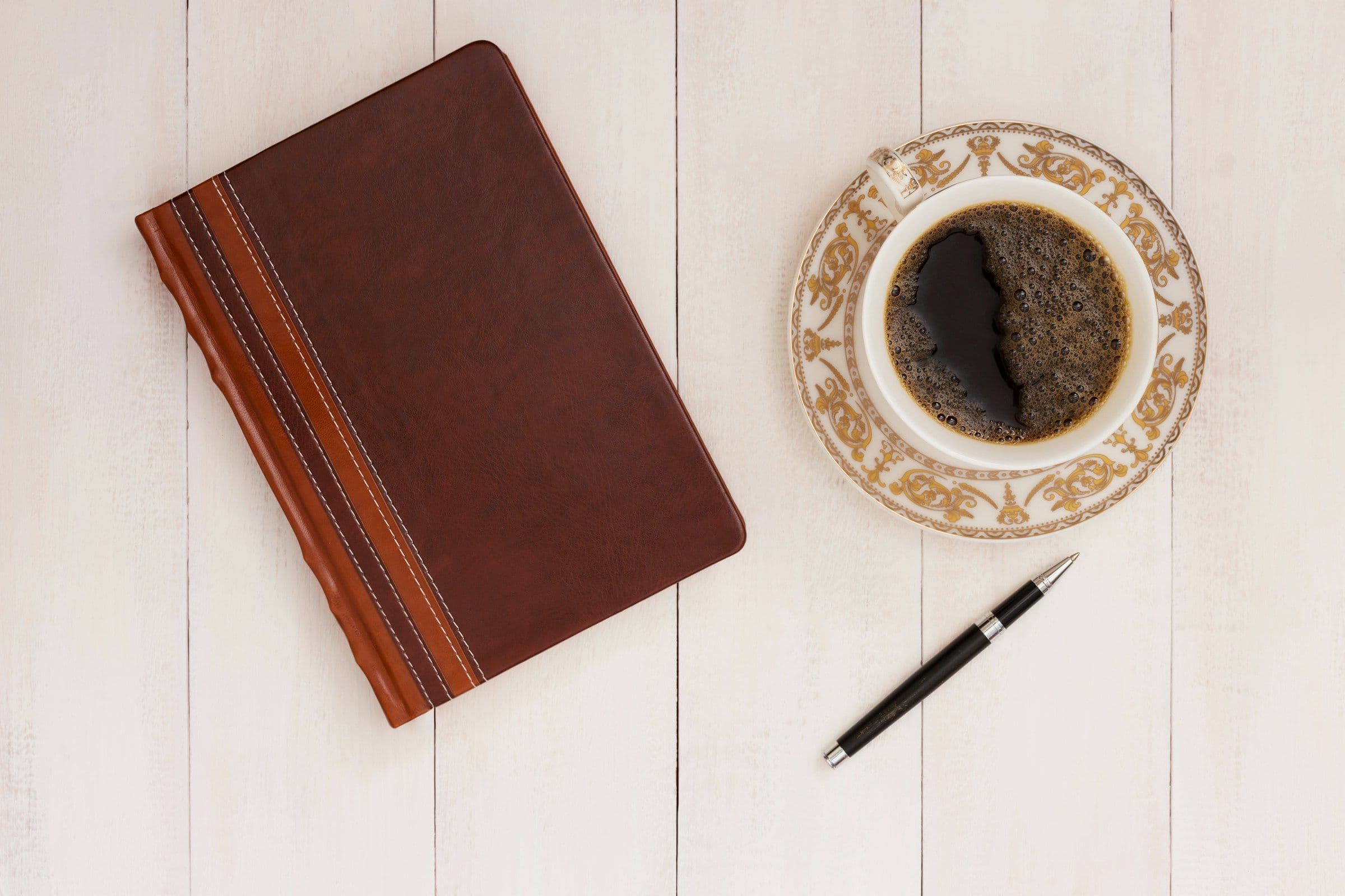 A leather notebook, pen, and cup of coffee on a wooden table